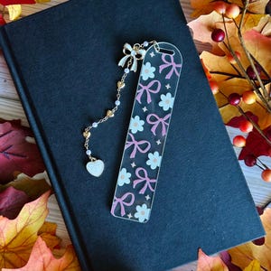 May include: A clear acrylic bookmark with a pattern of pink bows and white flowers, connected to a gold chain with a bow, hearts, and a heart-shaped charm. The bookmark rests on a closed black book.
