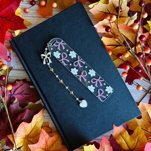 May include: A clear acrylic bookmark with a pink bow and flower pattern, accented with a gold chain and a heart charm. The bookmark rests on a closed black book, surrounded by autumn leaves.