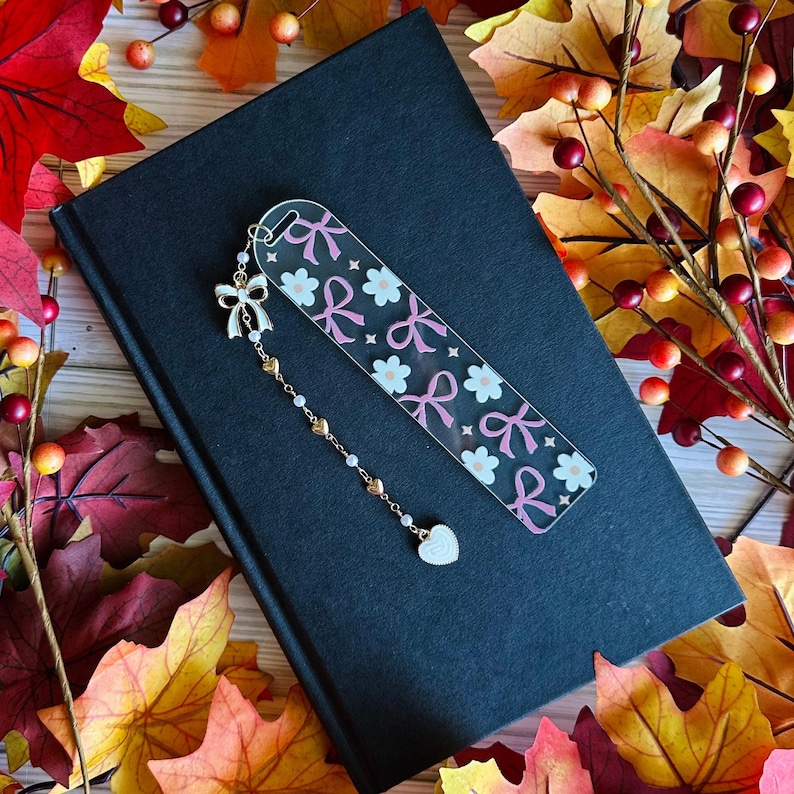 May include: A clear acrylic bookmark with a pink bow and flower pattern, accented with a gold chain and a heart charm. The bookmark rests on a closed black book, surrounded by autumn leaves.
