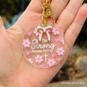 May include: Clear acrylic keychain with a gold clasp and a white bow. The keychain features pink flowers and hearts, with the words "She is Strong" and "Proverbs 31:25" in white. A white cross is also visible.
