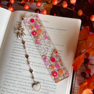 May include: A clear acrylic bookmark with a floral design in pink, orange, and yellow. The bookmark has a gold-colored chain with a white bow, pearls, and a heart charm. Text is visible on the bookmark.