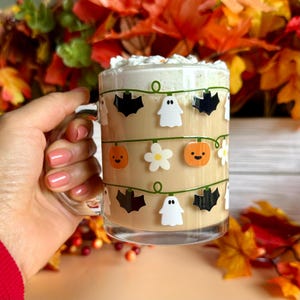 May include: A clear glass mug filled with a light brown beverage, topped with whipped cream. The mug is decorated with Halloween-themed stickers, including bats, ghosts, pumpkins, and flowers. The background features fall foliage.