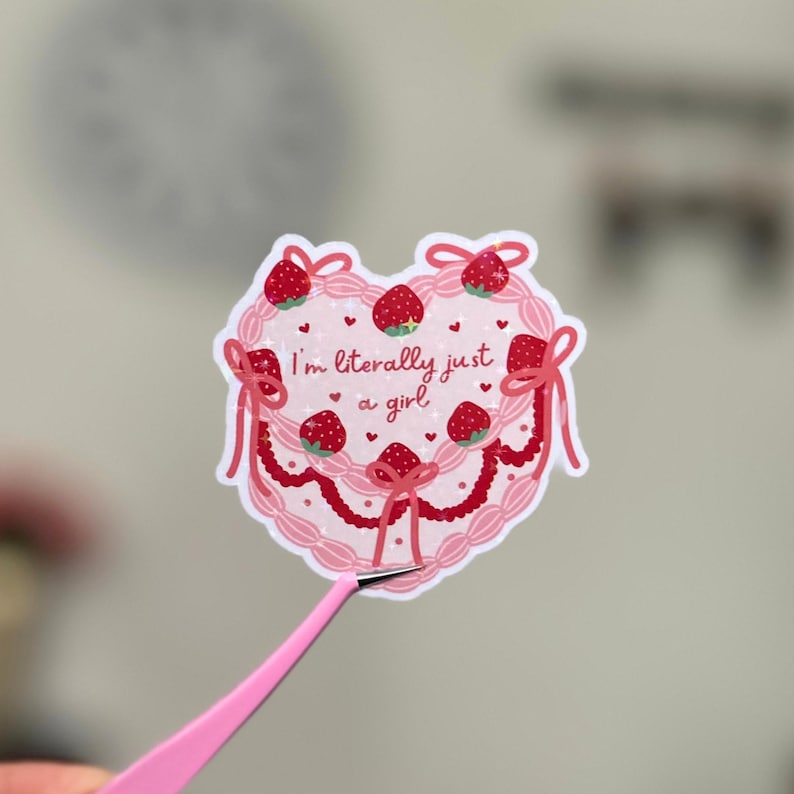 Puede incluir: Una pegatina rosa en forma de coraz&oacute;n con un dise&ntilde;o de pastel rosa y blanco. El pastel est&aacute; decorado con fresas y lazos. El texto "I'm literally just a girl" est&aacute; escrito en el pastel.
