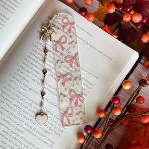 May include: A clear acrylic bookmark with a repeating pattern of pink bows and white flowers. The bookmark has a gold chain with heart and pearl charms, and a white heart charm at the end. The bookmark is resting on an open book.