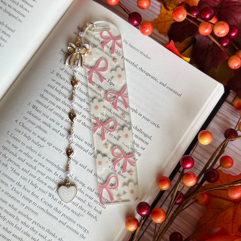 May include: A clear acrylic bookmark with a repeating pattern of pink bows and white flowers. The bookmark has a gold chain with heart and pearl charms, and a white heart charm at the end. The bookmark is resting on an open book.