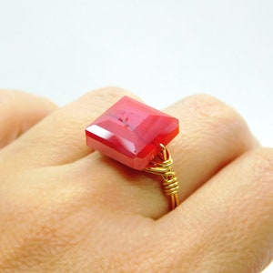 May include: A handmade ring featuring a faceted, square-cut, red gemstone. The gemstone is set on a gold-colored wire band, creating a unique and artistic design. The ring is shown on a hand.