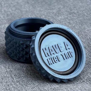 Peut inclure: Un récipient bleu imprimé en 3D avec un motif géométrique texturé. Le couvercle est ouvert, révélant l'intérieur et le texte "HAVE A NICE TRIP". Le récipient est posé sur une surface grise.