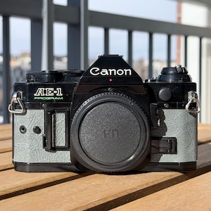Könnte beinhalten: Eine schwarze Canon AE-1 Program Kamera mit Objektivdeckel. Die Kamera hat einen grau strukturierten Griff und das Canon-Logo in Weiß. Der Text "AE-1 PROGRAM" ist grün. Die Kamera steht auf einer Holzoberfläche.