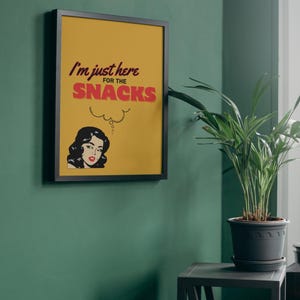 May include: A framed print with a yellow background and black frame. The print features a woman with black hair and a red dress, thinking about snacks. The text reads "I'm just here for the snacks".