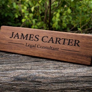 Puede incluir: Un letrero de escritorio rectangular de madera marrón oscuro con el nombre "JAMES CARTER" y el título "Legal Consultant" grabados en negro. El letrero está sobre una superficie de madera.
