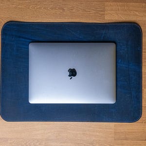 May include: A blue leather desk mat with a laptop computer on top. The laptop has a silver case and a black Apple logo.