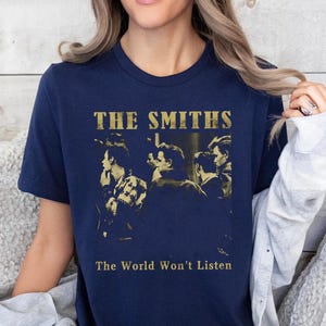 K&ouml;nnte beinhalten: Marineblaues T-Shirt mit dem Bandnamen "The Smiths" in goldfarbenen Buchstaben und einem Sepia-Bild der Band. Der Text "The World Won't Listen" ist darunter aufgedruckt. Das T-Shirt hat einen Rundhalsausschnitt.