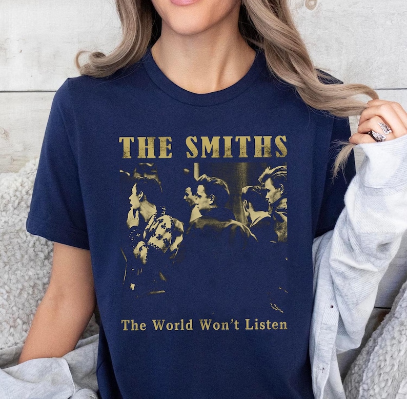 K&ouml;nnte beinhalten: Marineblaues T-Shirt mit dem Bandnamen "The Smiths" in goldfarbenen Buchstaben und einem Sepia-Bild der Band. Der Text "The World Won't Listen" ist darunter aufgedruckt. Das T-Shirt hat einen Rundhalsausschnitt.