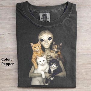 May include: A pepper-colored t-shirt featuring a graphic print of a grey alien holding three cats: one orange tabby, one white cat, and one black and white cat. The shirt has a crew neckline and a Comfort Colors brand label printed on the inside neck.