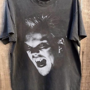 May include: Dark gray t-shirt featuring a black and white graphic of a vampire with fangs bared. The image is centered on the front of the shirt. The shirt is hanging on a wooden hanger.