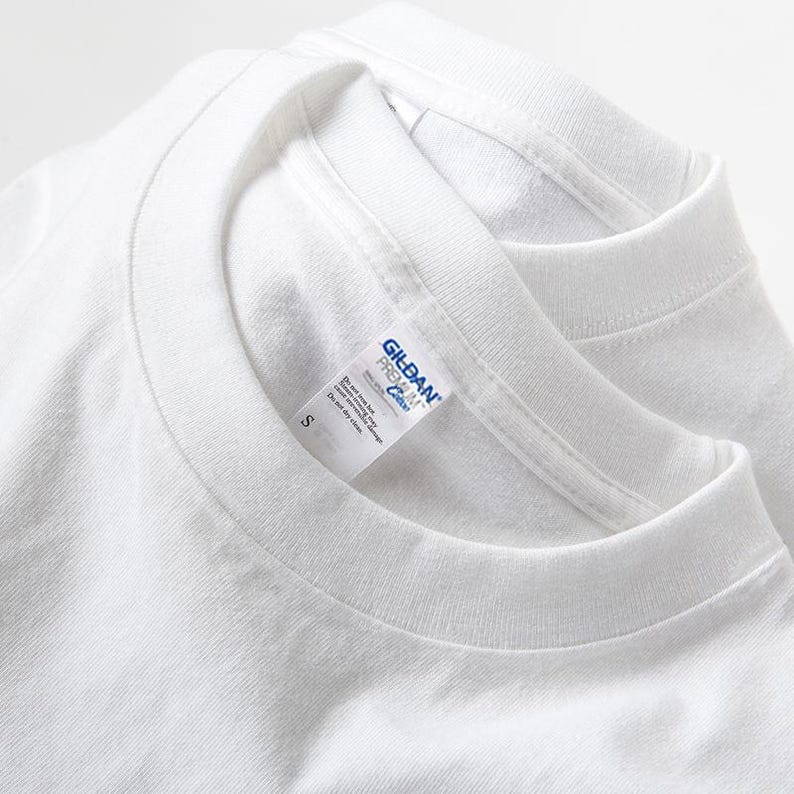 May include: Two white crew neck t-shirts are stacked, with the top shirt's collar and label visible. The label reads "Gildan Premium Cotton" and includes size "S". The shirts are made of a soft, plain white fabric.