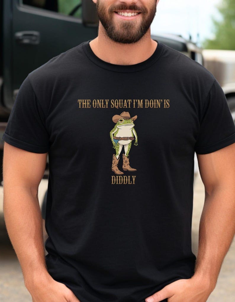 Puede incluir: Camiseta negra con una rana verde que lleva un sombrero de vaquero y botas. El texto de la camiseta dice "The only squat I'm doin' is diddly."