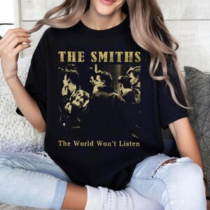K&ouml;nnte beinhalten: Schwarzes T-Shirt mit einem Sepia-Ton-Bild einer Band und dem goldenen Text "THE SMITHS" dar&uuml;ber und "The World Won't Listen" darunter. Das T-Shirt hat einen Rundhalsausschnitt.