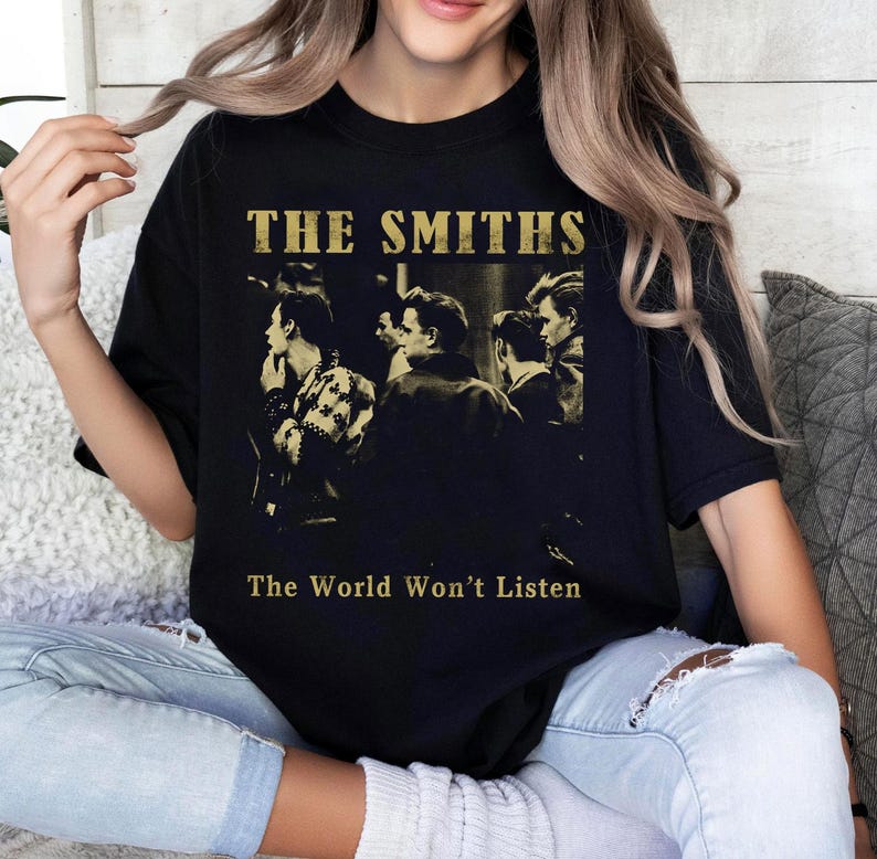 K&ouml;nnte beinhalten: Schwarzes T-Shirt mit einem Sepia-Ton-Bild einer Band und dem goldenen Text "THE SMITHS" dar&uuml;ber und "The World Won't Listen" darunter. Das T-Shirt hat einen Rundhalsausschnitt.
