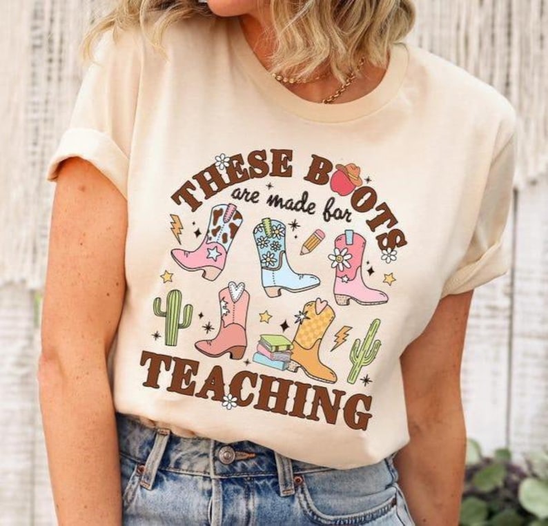 May include: Cream-colored t-shirt with a graphic design featuring colorful cowboy boots, cacti, and the text "These Boots are made for Teaching." The design includes stars, a pencil, and books.
