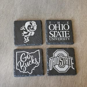 Può includere: Quattro sottobicchieri quadrati in ardesia con disegni incisi bianchi. I disegni includono il logo della Ohio State University, le parole "The Ohio State University", una mascotte di buckeye e le parole "Go Bucks" in una mappa dell'Ohio.
