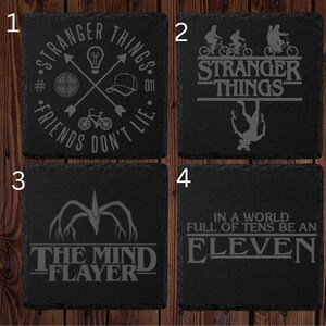 May include: Four black slate coasters with engraved designs. One coaster features the text "Stranger Things" with a lightbulb, bicycle, and hat. Another has the show's logo and upside-down silhouette. The third says "The Mind Flayer." The last says "In a world full of tens be an Eleven."