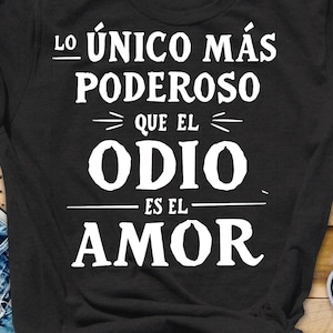 May include: Black t-shirt with white text that reads "Lo único más poderoso que el odio es el amor." The shirt is paired with blue jeans and beige sneakers on a wooden surface.