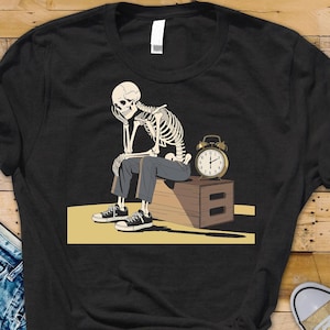 May include: Black t-shirt featuring a skeleton illustration. The skeleton is seated, resting its head on its hand, with an alarm clock nearby. The skeleton is wearing pants and sneakers. The design is on a yellow background.