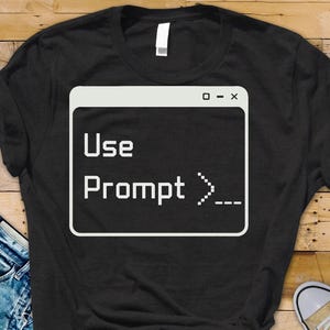 Könnte beinhalten: Schwarzes T-Shirt mit weißer Grafik eines Computer-Eingabeaufforderungsfensters. Der Text im Fenster lautet "Use Prompt >_-". Das Shirt besteht aus weichem Material und hat einen Rundhalsausschnitt.