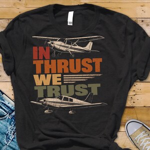 May include: Black t-shirt featuring two airplane illustrations and the text "IN THRUST WE TRUST" in orange, tan, and green. The shirt is paired with blue jeans and tan sneakers.