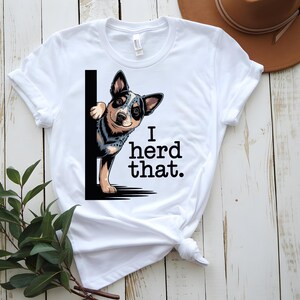 May include: White t-shirt featuring a cartoon dog peeking from behind a black vertical line. The dog has a blue, black, and tan coat. The text "I herd that." is printed in black below the dog.