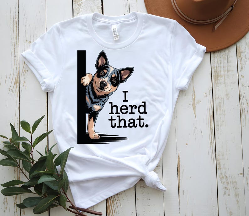 May include: White t-shirt featuring a cartoon dog peeking from behind a black vertical line. The dog has a blue, black, and tan coat. The text "I herd that." is printed in black below the dog.