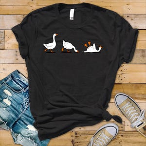 Puede incluir: Camiseta negra con un diseño humorístico de tres gansos blancos en varias poses. El primer ganso camina, el segundo se inclina y el tercero está boca abajo. Los gansos tienen patas y picos naranjas. La camiseta se combina con vaqueros azules y zapatillas de lona.