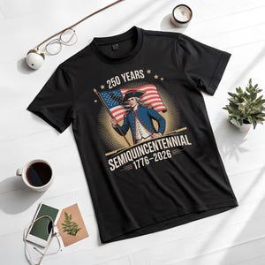 K&ouml;nnte beinhalten: Schwarzes T-Shirt mit einer Grafik eines Mannes in Kolonialkleidung, der eine amerikanische Flagge h&auml;lt. Das Design enth&auml;lt den Text "250 YEARS" und "SEMIQUINCENTENNIAL 1776-2026". Das Shirt wird auf einer wei&szlig;en Oberfl&auml;che mit Zubeh&ouml;r pr&auml;sentiert.