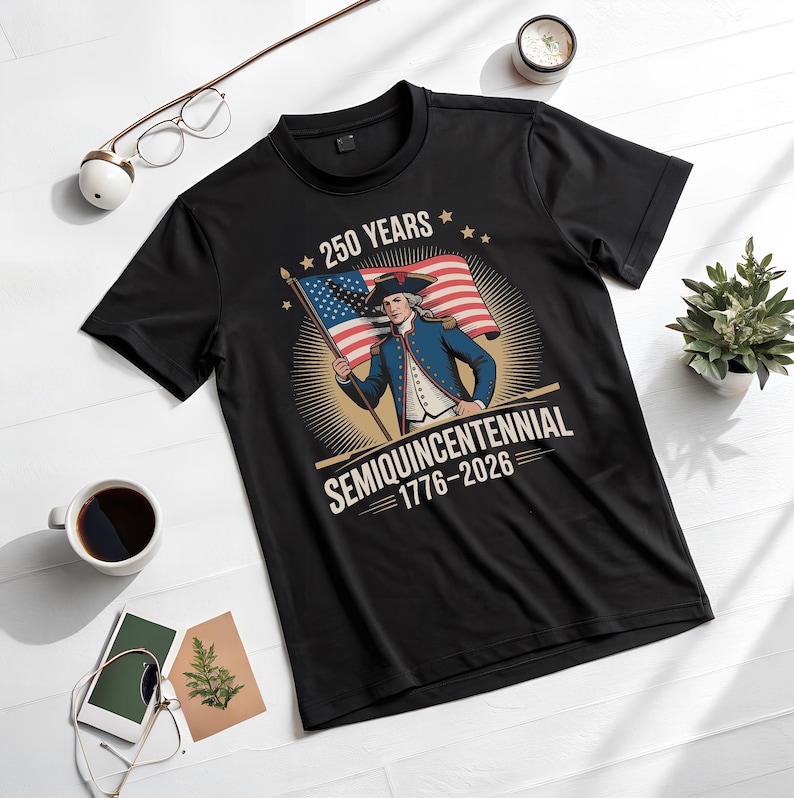 K&ouml;nnte beinhalten: Schwarzes T-Shirt mit einer Grafik eines Mannes in Kolonialkleidung, der eine amerikanische Flagge h&auml;lt. Das Design enth&auml;lt den Text "250 YEARS" und "SEMIQUINCENTENNIAL 1776-2026". Das Shirt wird auf einer wei&szlig;en Oberfl&auml;che mit Zubeh&ouml;r pr&auml;sentiert.