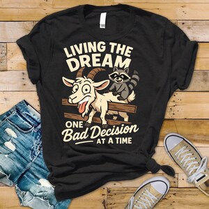 May include: Black t-shirt with a humorous graphic featuring a goat and raccoon on a fence, with the text "LIVING THE DREAM ONE BAD DECISION AT A TIME". The goat has a silly expression, and the raccoon is smiling.