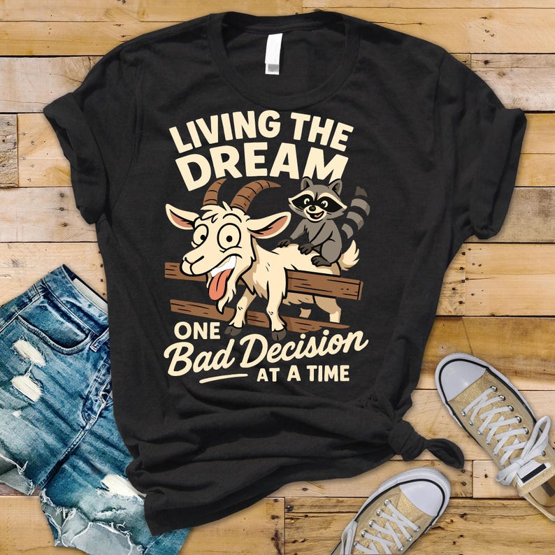 May include: Black t-shirt with a humorous graphic featuring a goat and raccoon on a fence, with the text "LIVING THE DREAM ONE BAD DECISION AT A TIME". The goat has a silly expression, and the raccoon is smiling.