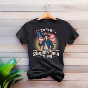 K&ouml;nnte beinhalten: Schwarzes T-Shirt mit einer Grafik eines Mannes in einem blauen Mantel, der eine amerikanische Flagge h&auml;lt. Der Text lautet "250 YEARS SEMIQUINCENTENNIAL 1776-2026" in Wei&szlig; und Gold. Das Design umfasst Sterne und einen Sonnenstrahl.