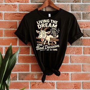 May include: Black t-shirt with a humorous graphic featuring a goat and raccoon on a fence. The text reads "LIVING THE DREAM" above and "ONE Bad Decision AT A TIME" below. The shirt is hanging on a wooden hanger against a brick wall.