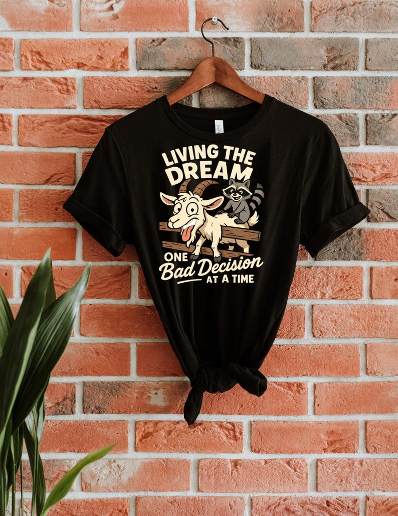 May include: Black t-shirt with a humorous graphic featuring a goat and raccoon on a fence. The text reads "LIVING THE DREAM" above and "ONE Bad Decision AT A TIME" below. The shirt is hanging on a wooden hanger against a brick wall.