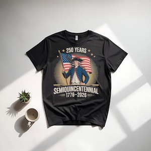 K&ouml;nnte beinhalten: Schwarzes T-Shirt mit einem Vintage-Grafikdesign eines Mannes in einem blauen Mantel, der eine amerikanische Flagge h&auml;lt. Der Text auf dem T-Shirt lautet "250 YEARS SEMIQUINCENTENNIAL 1776-2026". Das Design erinnert an ein historisches Ereignis.