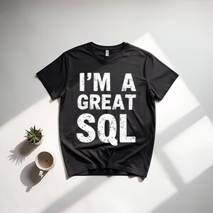 May include: Black crew neck t-shirt with the white text "I'M A GREAT SQL". The shirt is displayed on a white surface with a small potted plant and a mug.