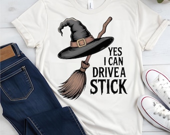 May include: Cream-colored t-shirt featuring a witch's hat and broom graphic. The hat is black with a brown band and buckle, and the broom is brown. The text "YES I CAN DRIVE A STICK" is printed in black.