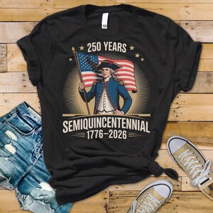 K&ouml;nnte beinhalten: Schwarzes T-Shirt mit einem Vintage-Grafik eines Mannes in einem blauen Mantel, der eine amerikanische Flagge h&auml;lt. Der Text "250 YEARS" steht &uuml;ber dem Bild und "SEMIQUINCENTENNIAL 1776-2026" darunter. Das Design feiert ein Jubil&auml;um.