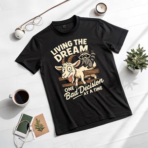 May include: Black t-shirt with a humorous graphic featuring a goat and raccoon on a wooden fence. The text reads "LIVING THE DREAM" above and "ONE BAD DECISION AT A TIME" below. The shirt is displayed on a white surface.