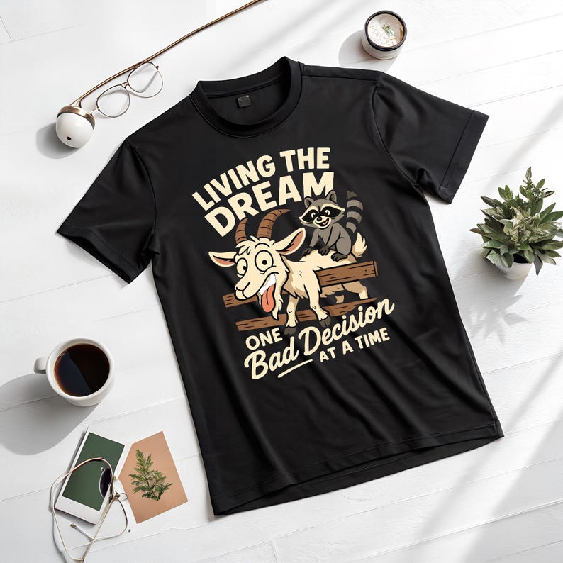 May include: Black t-shirt with a humorous graphic featuring a goat and raccoon on a wooden fence. The text reads "LIVING THE DREAM" above and "ONE BAD DECISION AT A TIME" below. The shirt is displayed on a white surface.