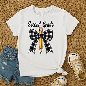 Può includere: T-shirt bianca con la scritta "Second Grade" in corsivo nero. Una matita gialla con gomma rosa è centrata, adornata da un fiocco a scacchi bianco e nero. La maglietta è annodata in basso.