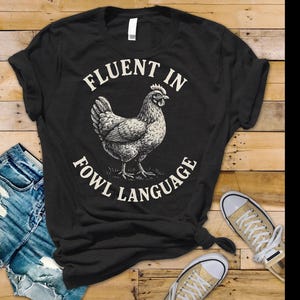 May include: Black t-shirt with the text "FLUENT IN FOWL LANGUAGE" surrounding a detailed illustration of a chicken. The shirt is paired with blue denim shorts and canvas sneakers on a wooden surface.