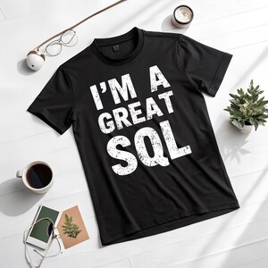 May include: Black crew neck t-shirt with the text "I'M A GREAT SQL" in white, distressed font. The shirt is displayed on a white surface, with a cup of coffee, glasses, and a small plant nearby.