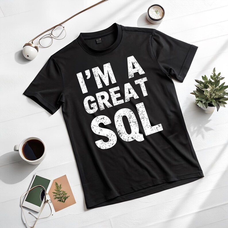 May include: Black crew neck t-shirt with the text "I'M A GREAT SQL" in white, distressed font. The shirt is displayed on a white surface, with a cup of coffee, glasses, and a small plant nearby.